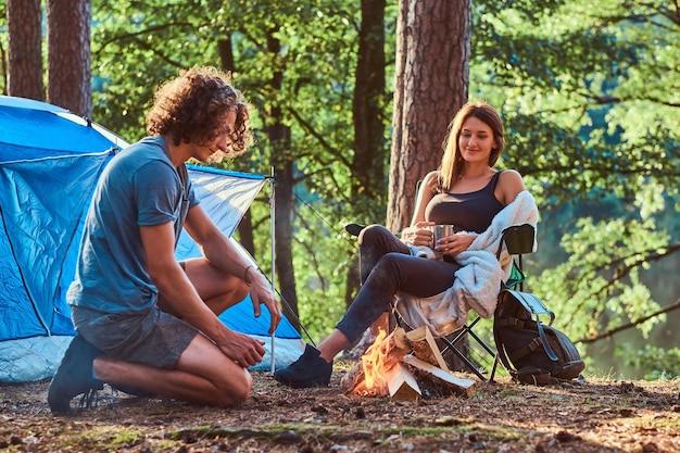 Ein paar junge Studenten chillen am Lagerfeuer im grünen Wald. Es gibt Zelt im Hintergrund.