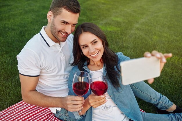 Ein Paar macht ein Selfie auf dem Smartphone, während es im Park mit einer Brille mit Rotwein klickt