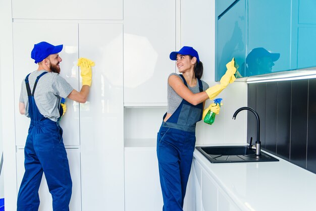 Ein paar professionelle Reinigungskräfte in blauer Uniform reinigen die Möbel mit Tüchern und Spray in der modernen Küche.