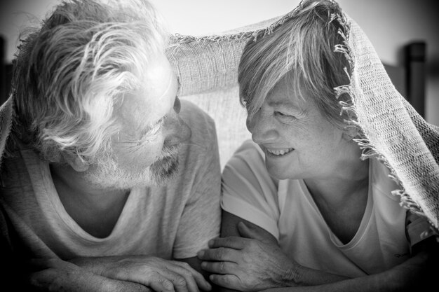 Foto ein paar senioren, die zwischendurch schauen und mit liebe und affekt unter der decke am bett lächeln - rentner und reife erwachsene, die morgens vor dem aufstehen im schlafzimmer heiraten