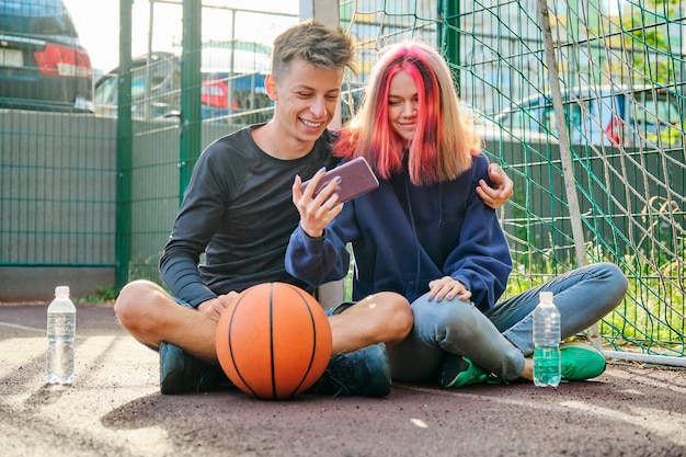 Ein paar Teenager auf dem Sportplatz mit Ball, Lifestyle, Jungen und Mädchen 16 Jahre alt reden, Wasser aus der Flasche trinken, die auf den Smartphone-Bildschirm schaut