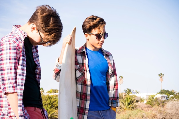 Ein paar Teenager mit Freundschaft am Strand mit Blick auf das Meer mit einem Surftisch, der dazwischen spricht - glücklicher Tag zusammen, bereit zum Surfen?