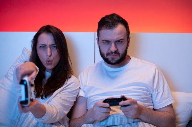 Ein paar verliebte Spieler spielen leidenschaftlich und emotional Videospiele im Bett mit Joysticks