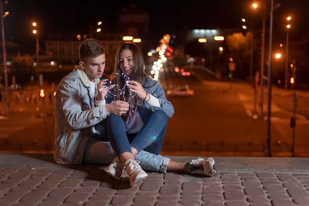 Ein paar verliebte Studenten sitzen am abendlichen Stadthintergrund mit einer Girlande in ihren Händen Nachtlichter der Stadt