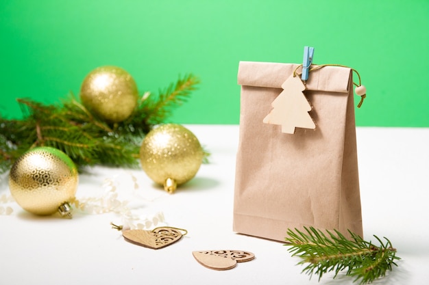 Ein Paket Öko-Papier verziert mit einem hölzernen Weihnachtsbaum auf einer Wäscheklammer, im Hintergrund ein Tannenzweig und goldene Weihnachtskugeln, Holzherzen, grüner Hintergrund