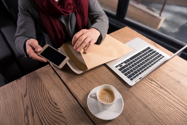 Ein Paket vorbereiten. Nahaufnahme eines modernen innovativen Smartphones, das von einem hart arbeitenden erfolgreichen Geschäftsmann beim Sitzen im Café mit Dokumenten in den Umschlag gesteckt wird