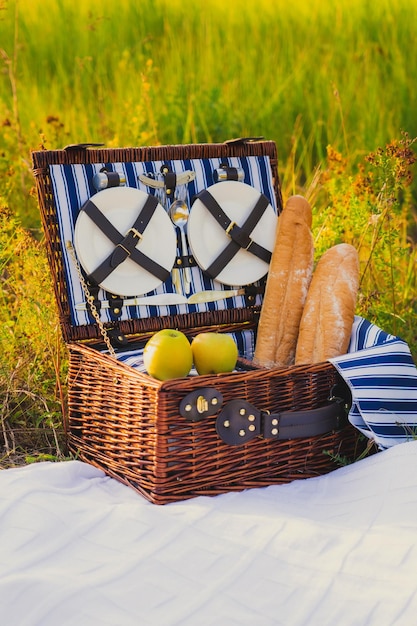 Ein Picknickplatz, ein Weidenkoffer für Picknickgeschirr mit grünen Äpfeln und einem Baguette steht auf einer weißen Decke.