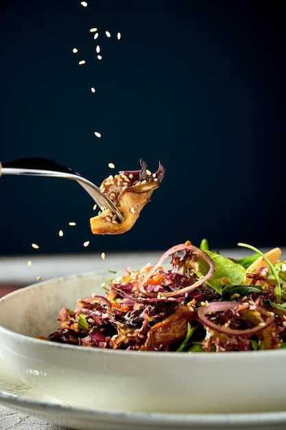 Ein Pilz auf einer Gabel wird mit Sesam vor blauem Hintergrund bestreut Vegetarischer Salat in einer Schüssel