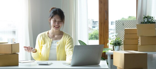 Ein Porträt einer asiatischen E-Commerce-Mitarbeiterin, die im Büro voller Pakete auf dem Tisch sitzt und einen Laptop und Taschenrechner für KMU-E-Commerce-Technologie und Liefergeschäft verwendet