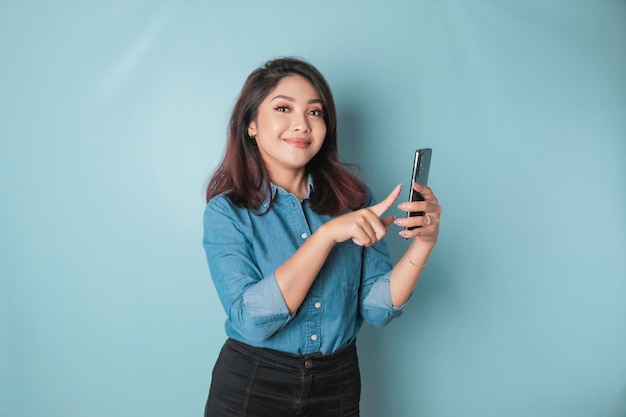 Ein Porträt einer glücklichen asiatischen Frau lächelt und hält ihr Smartphone in einem blauen Hemd, das durch einen blauen Hintergrund isoliert ist