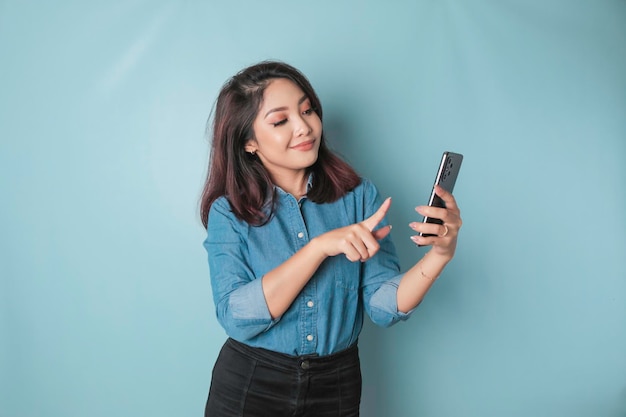 Ein Porträt einer glücklichen asiatischen Frau lächelt und hält ihr Smartphone in einem blauen Hemd, das durch einen blauen Hintergrund isoliert ist