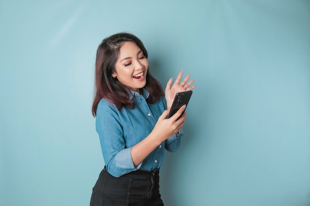 Ein Porträt einer glücklichen asiatischen Frau lächelt und hält ihr Smartphone in einem blauen Hemd, das durch einen blauen Hintergrund isoliert ist