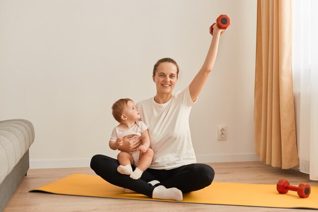 Ein Porträt einer schönen jungen Frau in einer Sportuniform, die mit einem Kind in ihren Armen posiert, das Bizeps und Trizeps trainiert und eine rote Hantel mit Blick auf die Kamera hebt Fitness zu Hause