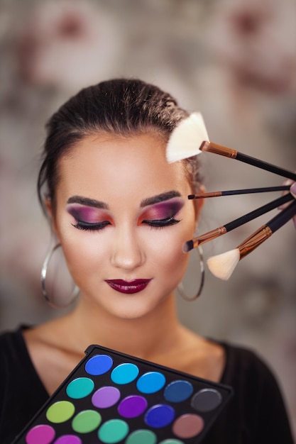 Ein Porträt einer süßen jungen Frau mit perfektem Make-up, umgeben von vielen Make-up-Pinseln und Paletten, posiert im Make-up-Studio.