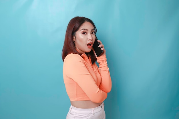 Ein Porträt einer überraschten jungen asiatischen Frau, die einen Anruf auf blauem Hintergrund hat