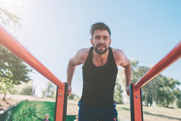 Ein Porträt eines fokussierten muskulösen bärtigen Mannes in schwarzer Trainingskleidung, der Dips auf Barren macht. Mans Fitness mit blauem Himmel im Hintergrund und offenem Raum um ihn herum. Sport und Crossfit.