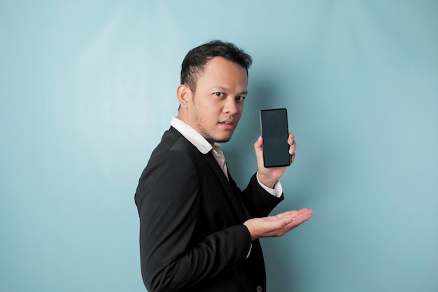 Ein Porträt eines glücklichen asiatischen Geschäftsmannes lächelt und hält sein Smartphone in der Hand, das einen Kopierbereich auf dem Bildschirm des it39 zeigt, der einen schwarzen Anzug trägt, der durch einen blauen Hintergrund isoliert ist