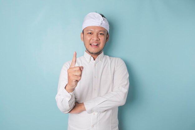 Ein Porträt eines glücklichen balinesischen Mannes lächelt und trägt Udeng oder traditionelles Stirnband und ein weißes Hemd, das von einem blauen Hintergrund isoliert wird