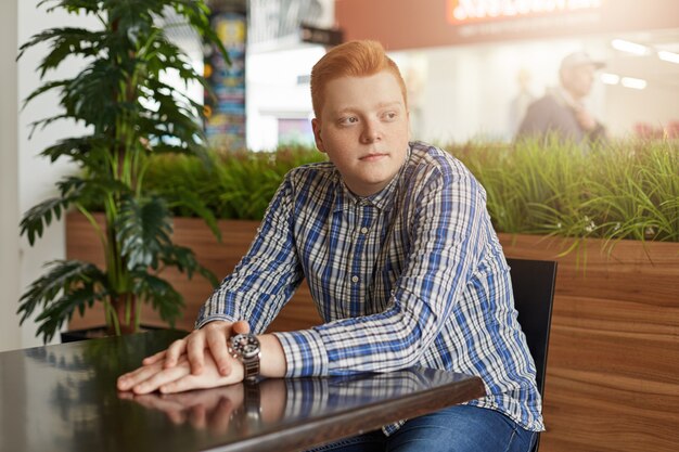 Ein Porträt eines gutaussehenden Mannes mit rotem Haar, der kariertes Hemd und Jeans trägt, die am Holztisch nahe grüner Handfläche sitzen