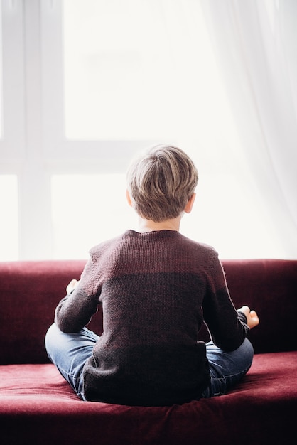 Ein Porträt eines Jungen, der auf einem Sofa in Lotus-Pose-Ansicht von seinem Rücken sitzt, weicher Fokus