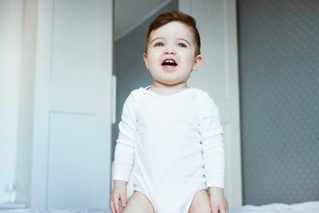 Ein Porträt eines niedlichen Kleinkindbabys in einem weißen Bodysuit, der auf Bett sitzt und glückliches Babygesicht im Schlafzimmer zu Hause lacht