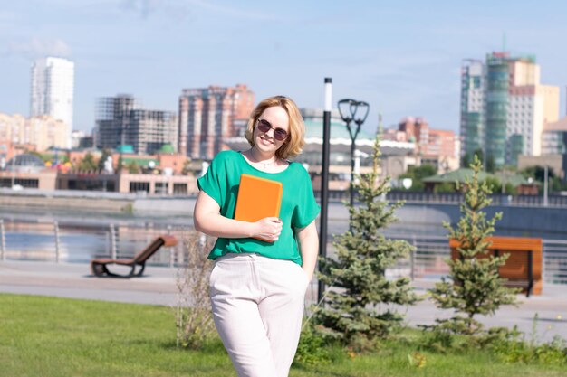 Ein Porträt eines Studentenmädchens, das mit Sonnenbrille lächelt und ein digitales Tablet in den Händen auf dem Stadtdamm hält