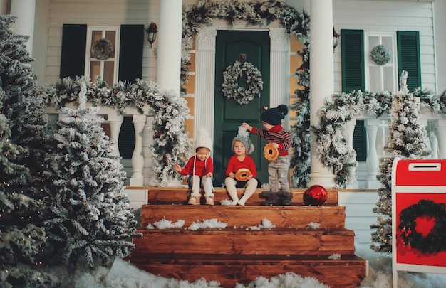 Ein Porträt von Kindern, die zusammen auf der Veranda ihres Hauses sitzen, essen Bagels. Frohe Weihnachten, Frohes neues Jahr. Hof mit einem Weihnachtsbaum, Lichtern und Dekorationen. Wunderzeit.