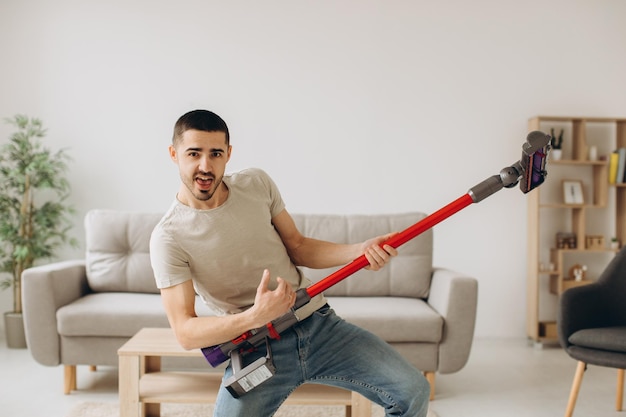 Ein positiver junger Mann putzt den Raum und hält einen Staubsauger wie eine Gitarre