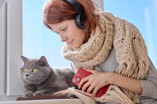 Ein Preteen-Mädchen in einem gestrickten Wollschal mit einer niedlichen grauen Katze in der Nähe des Winterfensters Ein Kind in Kopfhörern mit Telefon zusammen mit einem Haustier an einem winterlichen sonnigen Tag