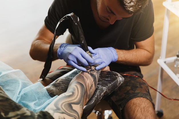 Ein professioneller Tätowierer, der maschinell mit schwarzer Tinte am Arm eines jungen Mannes tätowiert