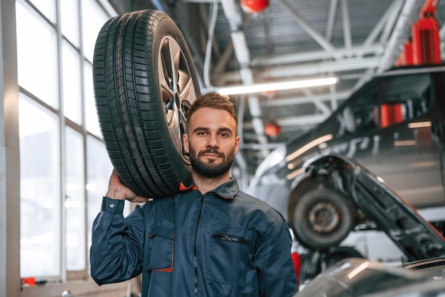 Ein Reifen in der Hand, ein Autoreparaturmann ist in der Garage mit einem Auto