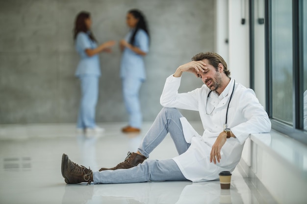 Ein reifer Arzt sieht besorgt aus, während er während der Covid-19-Pandemie eine schnelle Kaffeepause macht und auf dem Boden neben einem Fenster in einem leeren Krankenhausflur sitzt.