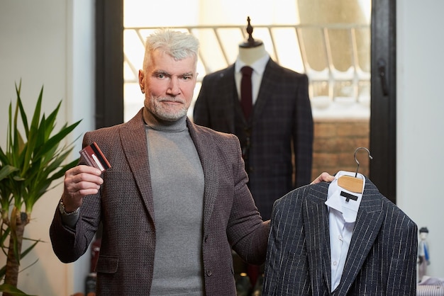 Ein reifer Mann mit grauen Haaren und sportlichem Körperbau posiert mit Wollanzug und Kreditkarte in einem Bekleidungsgeschäft Ein männlicher Kunde mit Bart trägt in einer Boutique einen Anzug