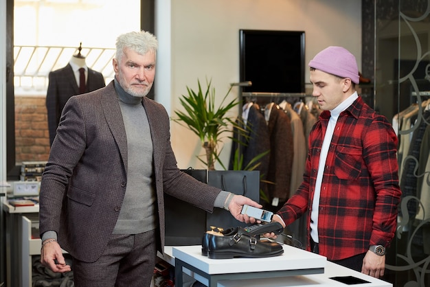 Ein reifer Mann mit grauen Haaren und sportlichem Körperbau wendet ein Smartphone an einem Point-of-Sale-Terminal in einem Bekleidungsgeschäft an. Ein männlicher Kunde mit Bart bezahlt in einer Boutique an eine Verkäuferin.