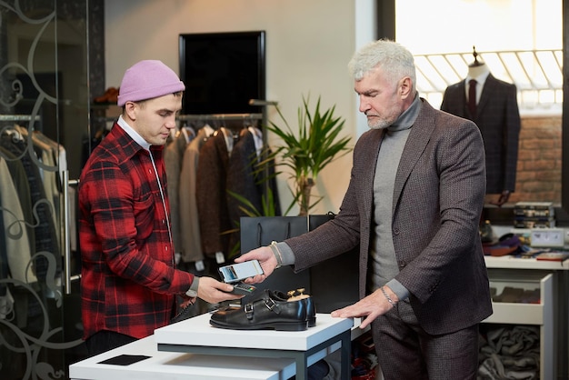 Ein reifer Mann mit grauen Haaren und sportlicher Statur hält ein Handy an einem Kassenterminal in einem Bekleidungsgeschäft an Ein männlicher Kunde mit Bart bezahlt bei einer Verkäuferin in einer Boutique