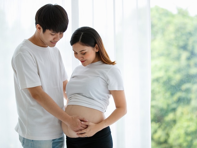 Ein reizendes junges asiatisches Paar, das zusammen steht und den Bauch einer schwangeren Frau mit Liebe und Sorgfalt lächelt und berührt. Glückliches und gesundes Familienkonzept.