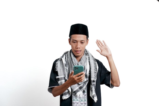 Ein religiöser muslimischer asiatischer Mann, der eine muslimische Turban-Kleidung und eine Mütze trägt, die ein Smartphone wie he39s bei einem Videoanruf hält und schockiert ist, isoliert auf weißem Hintergrund