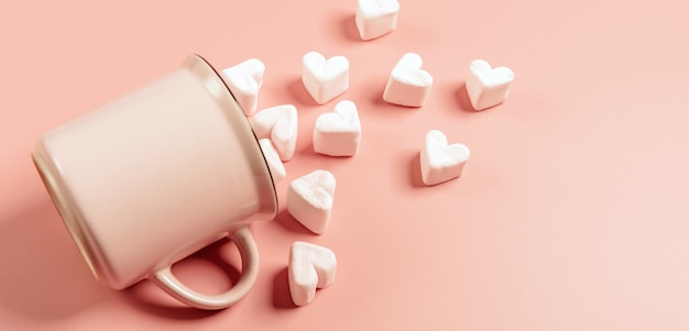 Ein rosa Becher liegt auf der Seite vor einer rosa Oberfläche, hellrosa Marshmallows in Form von Herzen