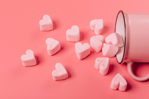 Ein rosa Becher liegt auf der Seite vor einer rosa Oberfläche, hellrosa Marshmallows in Form von Herzen