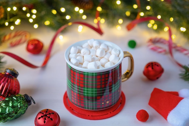 Ein rot karierter Becher mit Marshmallows auf einem Hintergrund aus Weihnachtsdekorationen und Tannenzweigen mit beleuchteter Girlande