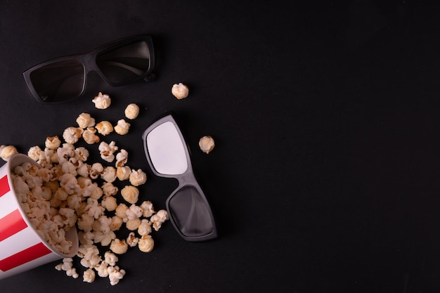 Ein rot-weißer, verstreuter Eimer Karamell-Popcorn und Gläser für 3D-Filme auf schwarzem Hintergrund Draufsicht Das Konzept eines Banners oder Werbung für ein Kino Platz für Text