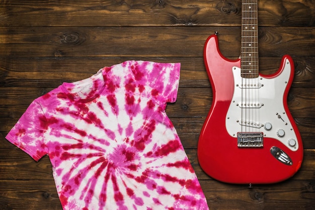 Ein rot-weißes Tie-Dye-T-Shirt und eine rot-weiße Gitarre auf einem hölzernen Hintergrund. Färben von Stoff im Tie-Dye-Stil. Flach liegen.