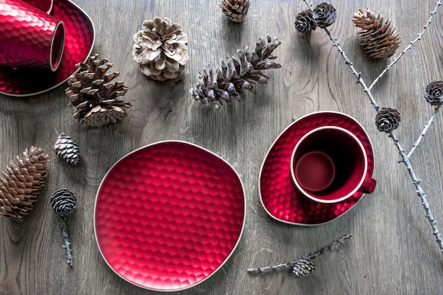 Ein rotes Geschirr: eine Teetasse, eine Untertasse, ein Teller und verschiedene Zapfen auf einem hölzernen Hintergrund. Weihnachtstischdekoration, magische Feiertage.