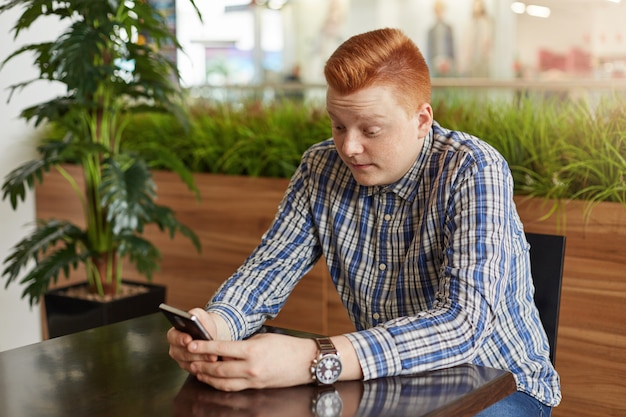 Ein rothaariger Mann, der elegantes Hemd sitzt, das am hölzernen Tisch hält, der Handy hält, das beim Lesen von Nachrichten online überrascht wird