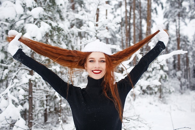 Ein rothaariges Mädchen mit langen Haaren in einer weißen Mütze steht und lächelt in einem schneebedeckten Winterwald. Großes Porträt eines rothaarigen Mädchens.