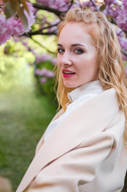 Ein rothaariges Mädchen mit rosa Lippenstift steht in der Nähe eines rosa blühenden Baumes, schaut in den Rahmen und lächelt.