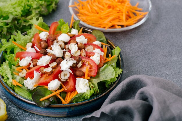 Ein Salat aus frischen Salatblättern, Tomaten, Mozzarella, Karotten und Haselnüssen mit Olivenöl und Zitronensaft in einer Keramikplatte auf grauem Hintergrund, Platz für Text.