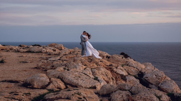 Ein sanftes junges Hochzeitspaar Braut und Bräutigam gehen und umarmen sich am Abend an einem felsigen Strand in der Nähe des Meeres auf Zypern Schöne Hochzeits-Fotoshooting Hochzeit für zwei Mal für zwei