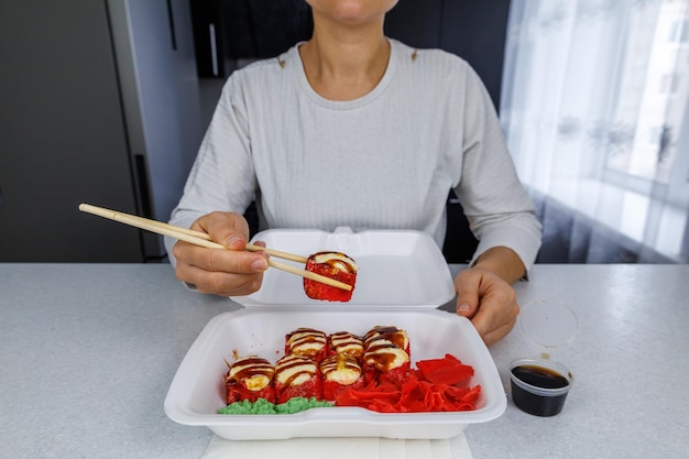 Ein Satz japanischer gebackener Brötchen in einer Plastikbox auf einem hellen Tisch zu Hause in der Küche Das Mädchen hält Essen mit Essstäbchen in der Hand Sushi mit schneller Lieferung in einem weißen Behälter