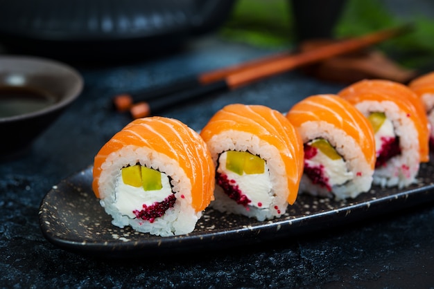 Ein Satz Sushi-Rollen Philadelphia mit rotem Fisch und Frischkäse liegt in einem Tellerboot. Sushi rollt auf einem schwarzen Hintergrund mit einer Teekanne des chinesischen Tees.